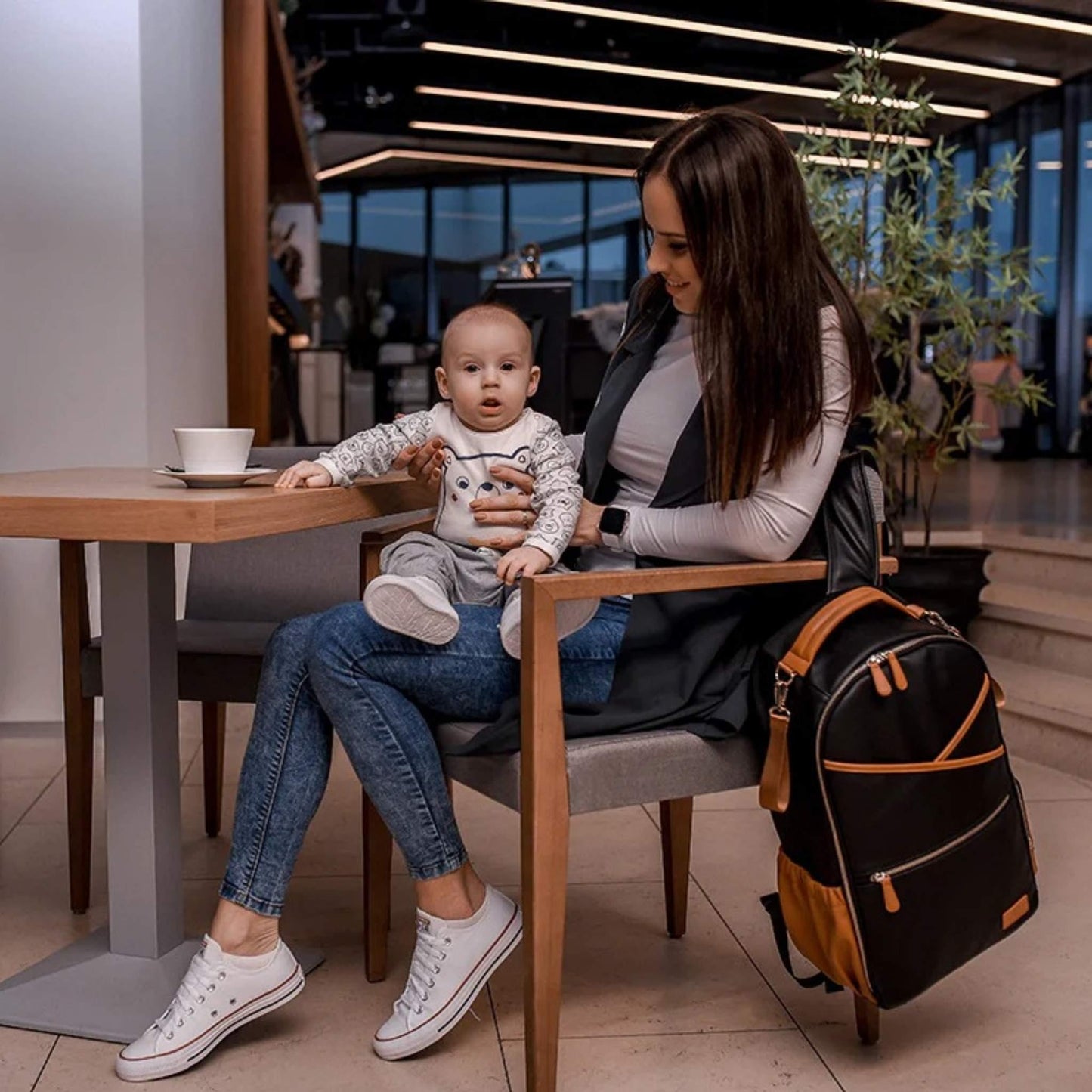 Woman sitting at a table with a child, surrounded by a backpack and a cup.