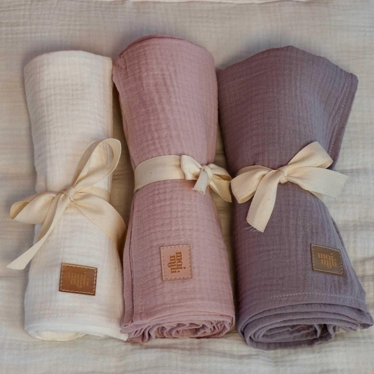 Three folded fabric swaddles in white, pink, and purple with visible brand labels on a textured surface.