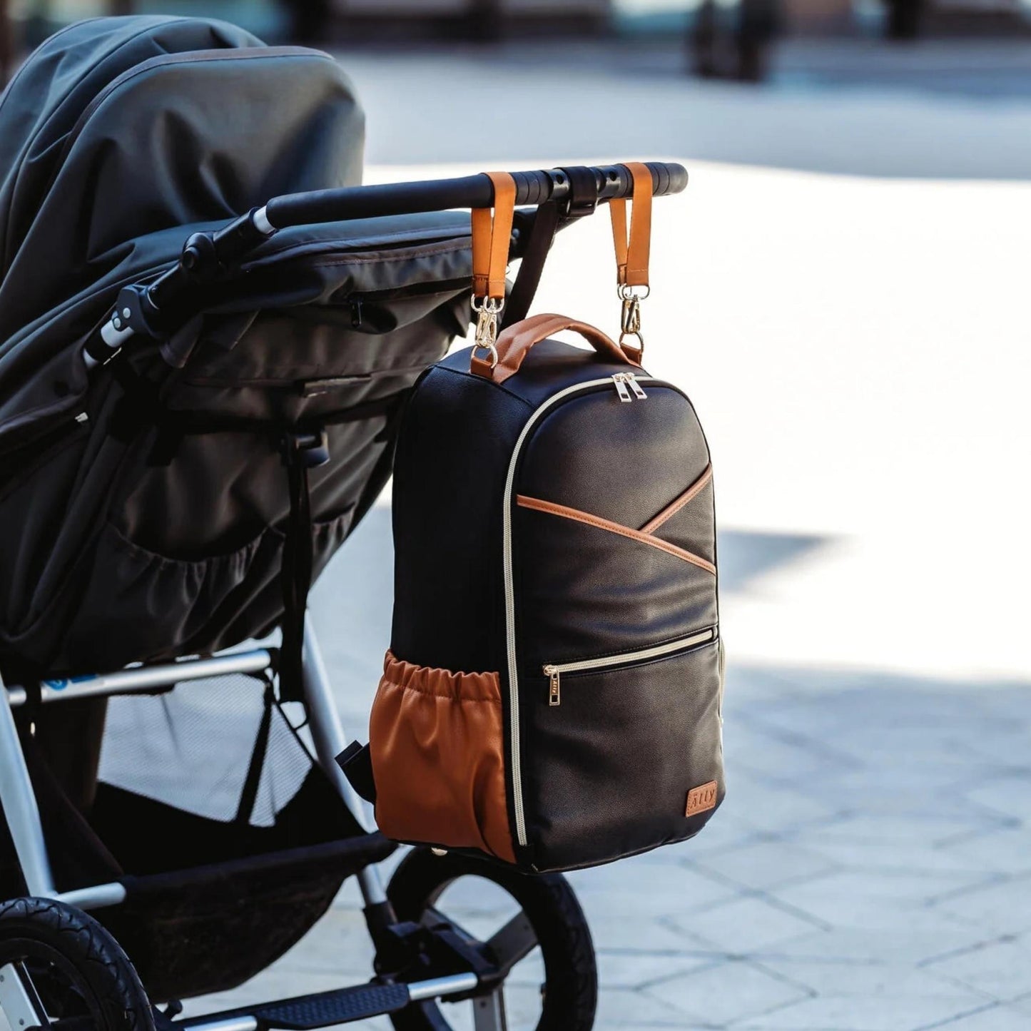 Black diaper bag with brown accents attached to a stroller on a blurred street background