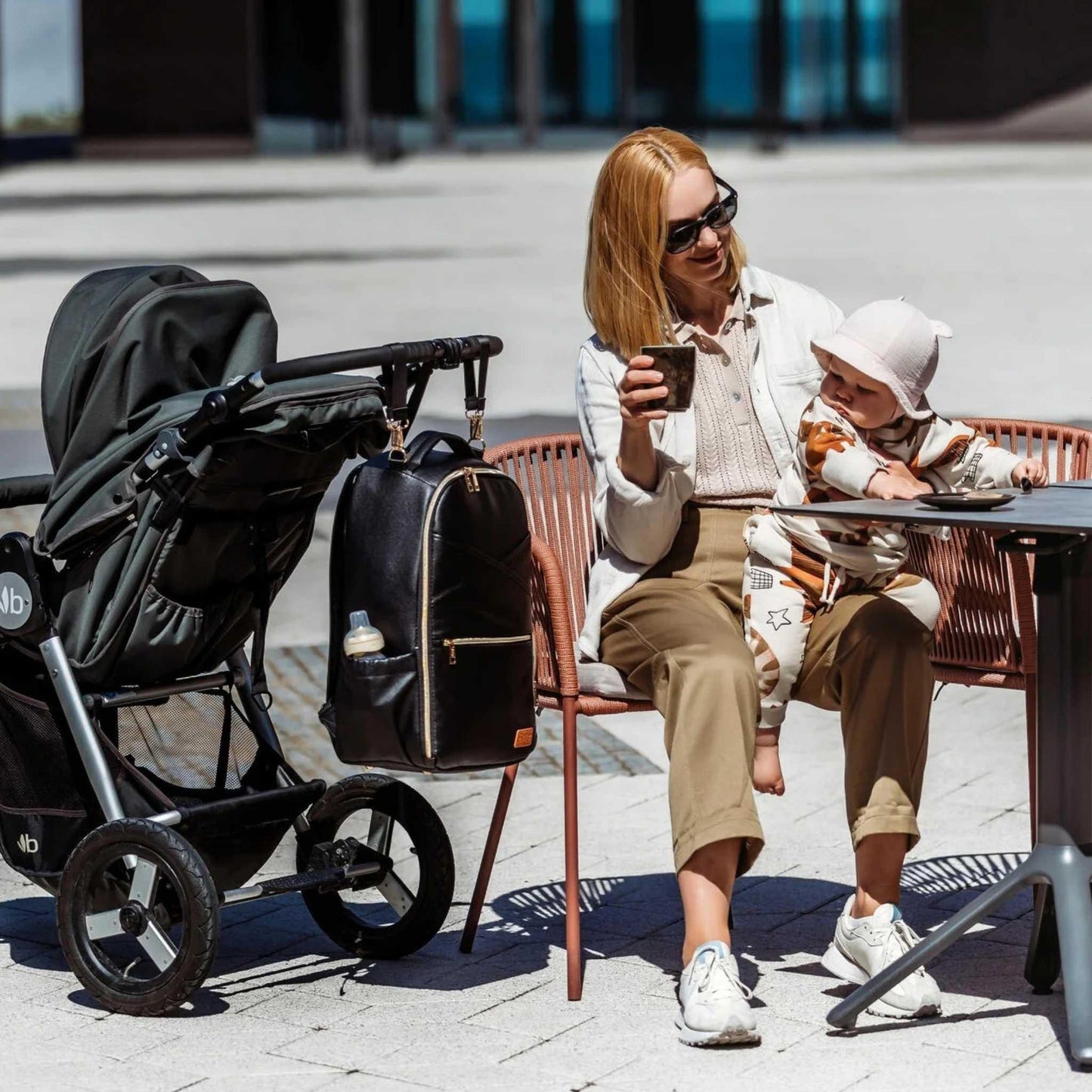 Woman sitting at an outdoor table with a baby in a stroller and a backpack next to her.