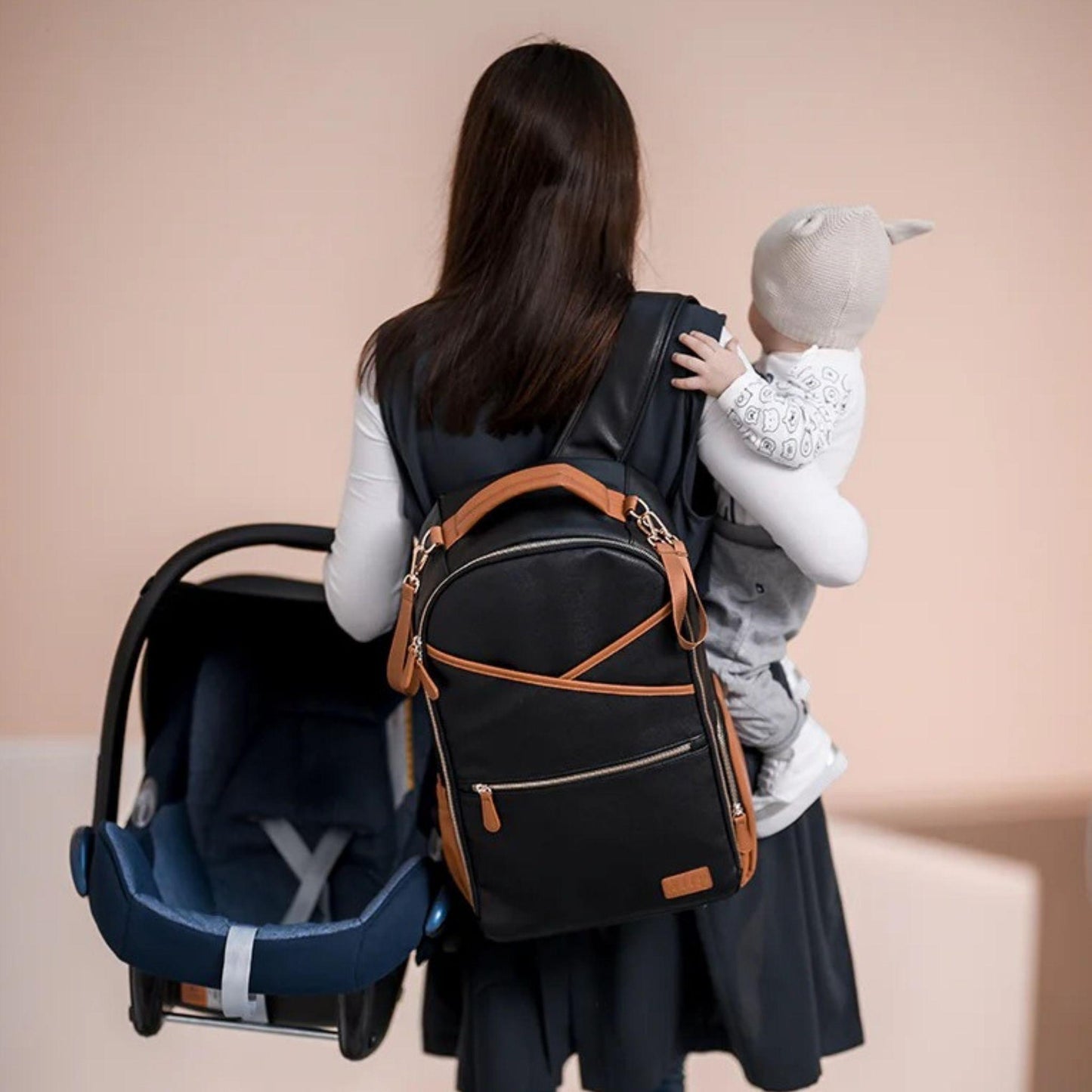 Woman carrying a baby and a black diaper bag with brown straps on a beige background