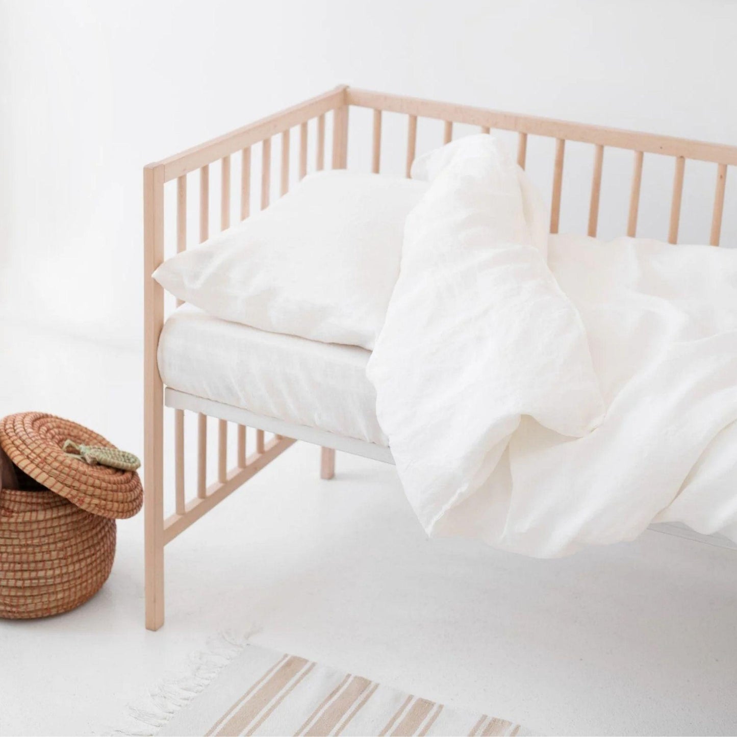 Nursery crib with white bedding and a woven basket on a light wooden floor.
