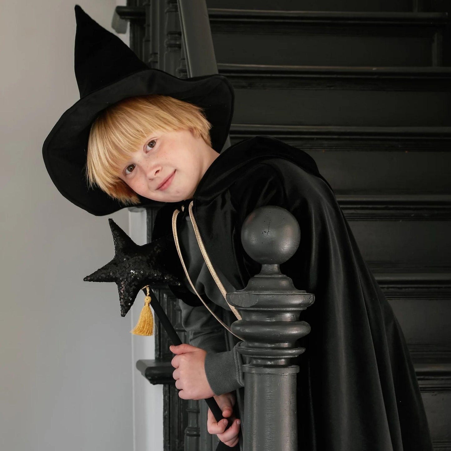 Child in a witch costume with a black hat and cape, standing on a staircase.
