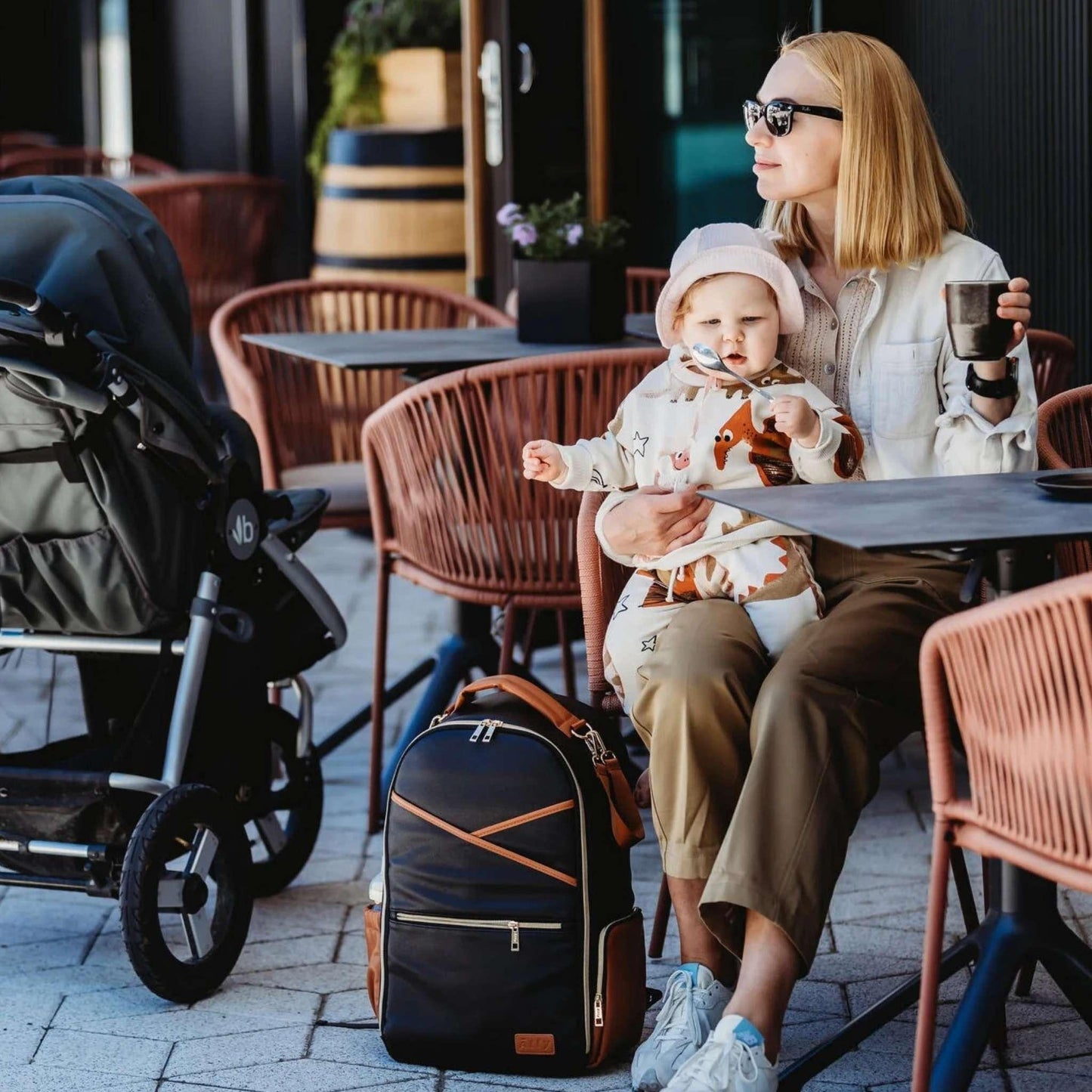 Woman with a child sitting at an outdoor cafe table with a stroller and diaper bag nearby.