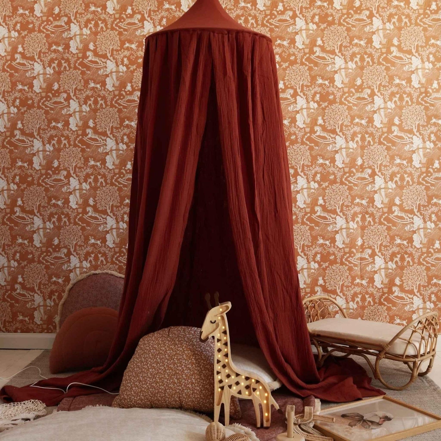 Red canopy over a child's bed with patterned wallpaper and toys in a nursery.