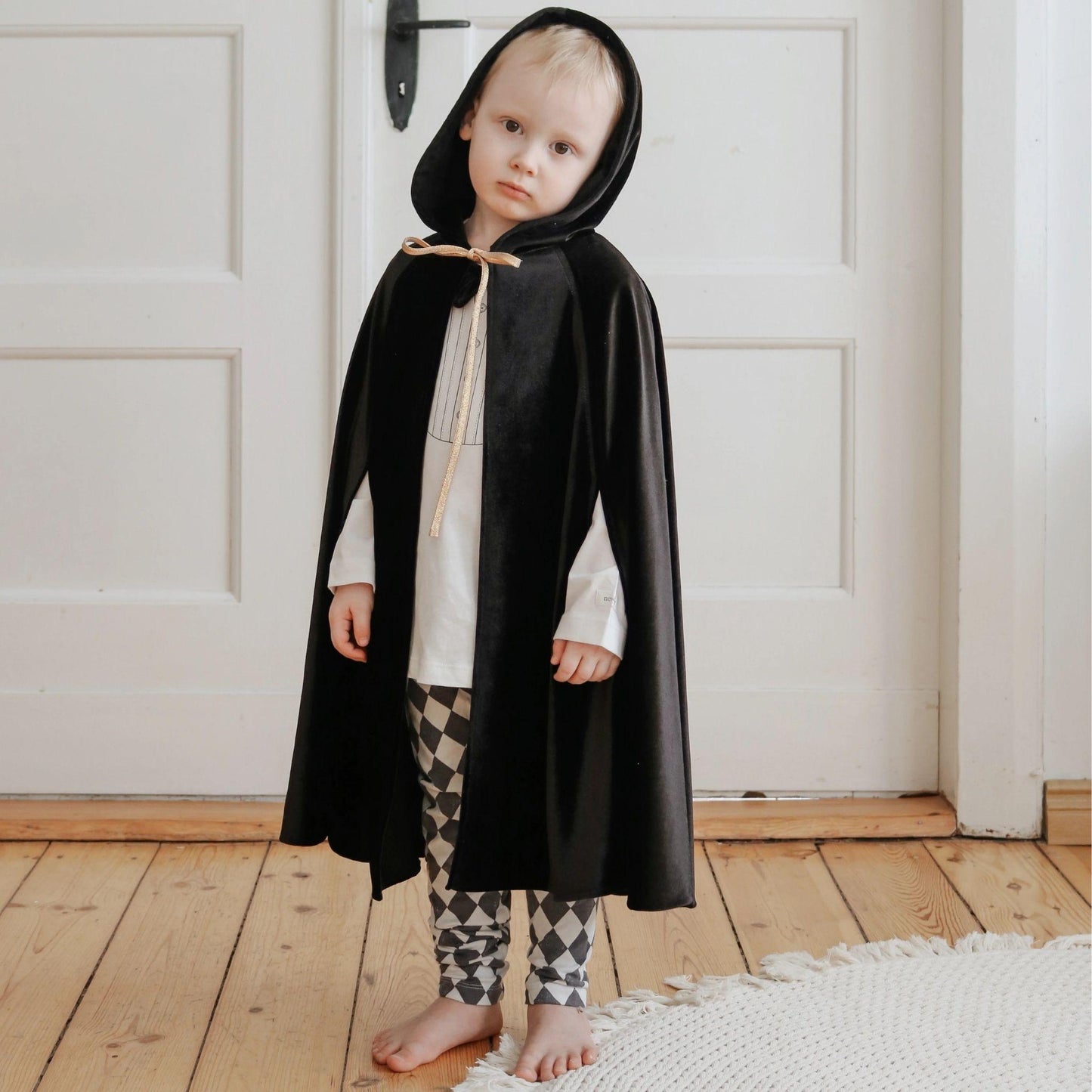 Child wearing a black hooded cloak indoors on a wooden floor.