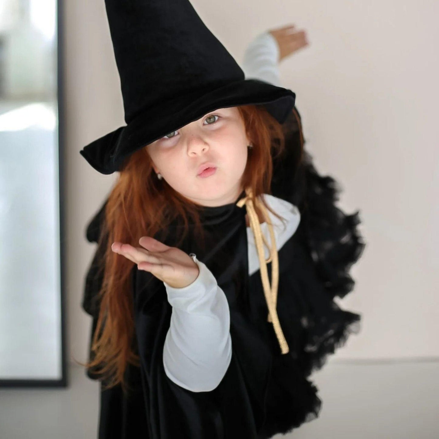 Child in a witch costume with a black hat and cape, blowing a kiss.