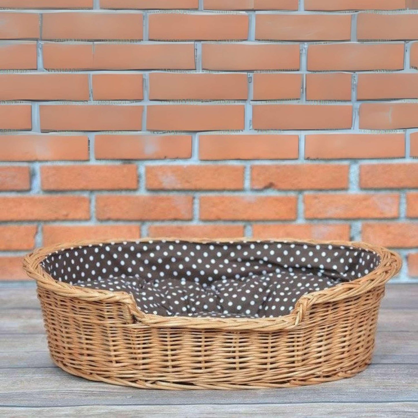 Wicker pet bed with a brown and white polka dot cushion against a brick wall.