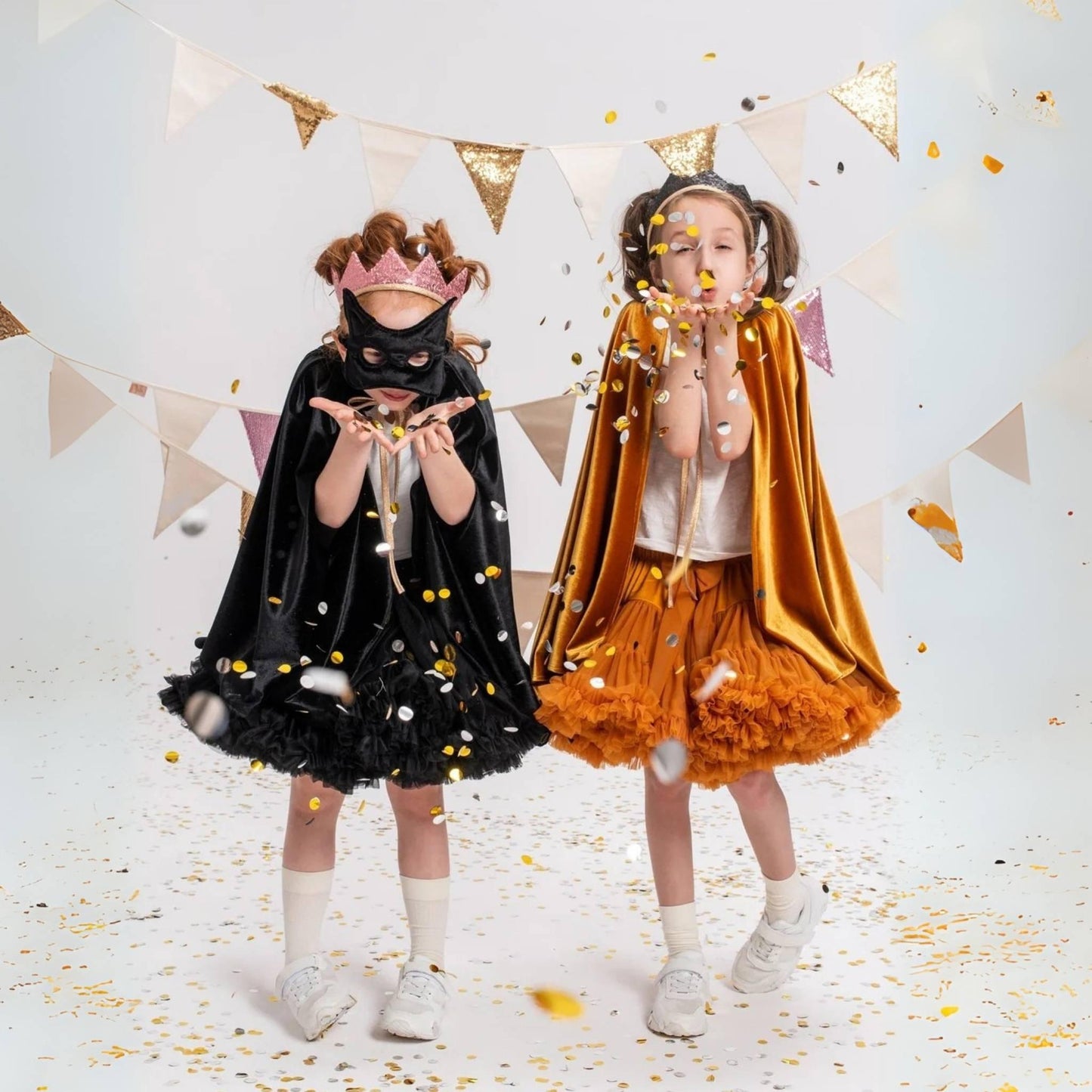 Two children in superhero costumes with masks and capes, surrounded by confetti and party decorations.