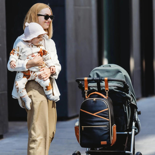 Woman holding a baby and pushing a stroller with a backpack on a city street.
