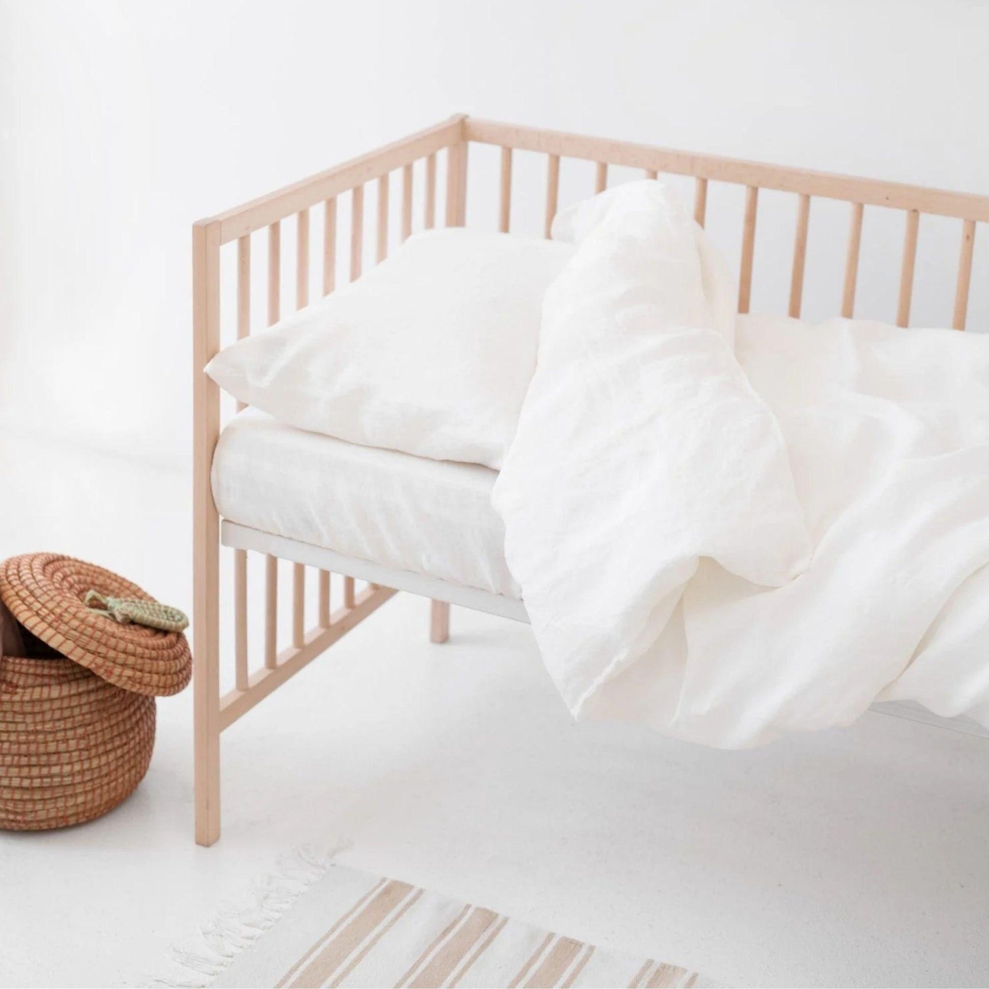 Nursery crib with white bedding and a woven basket on a light wooden floor.