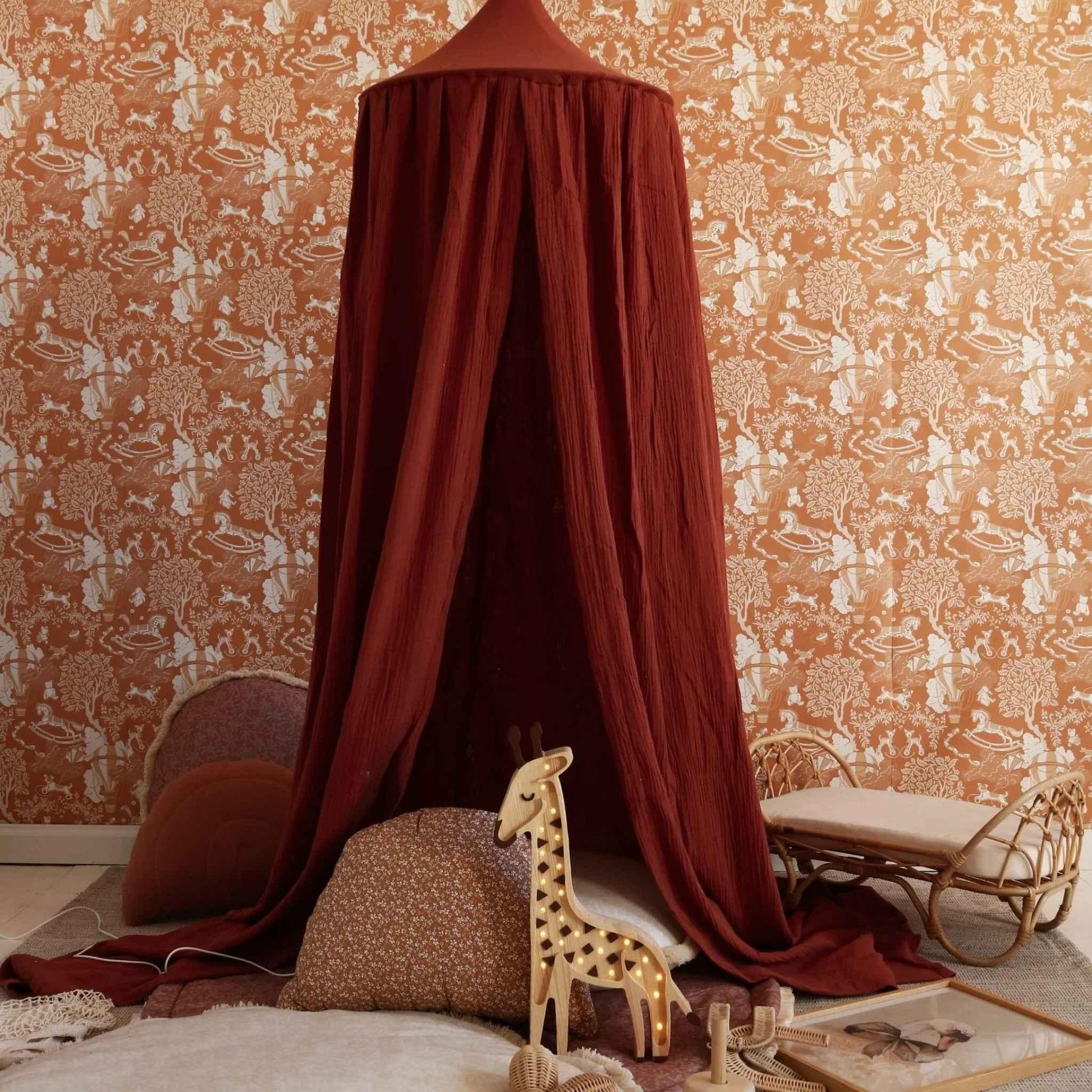 Red canopy over a child's bed with patterned wallpaper and toys in a nursery.
