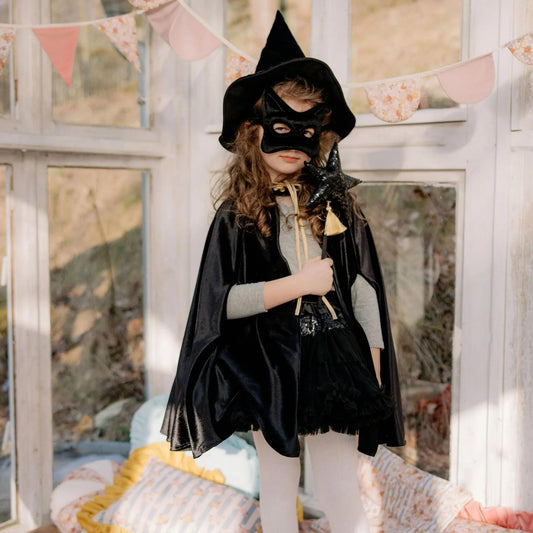 Child in a witch costume with a black hat and cape indoors.