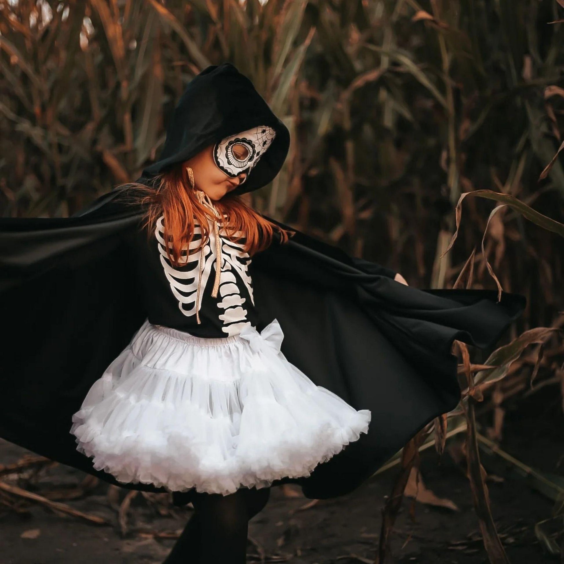 Child in a Halloween costume with a black cape and skeleton design, standing in a field.
