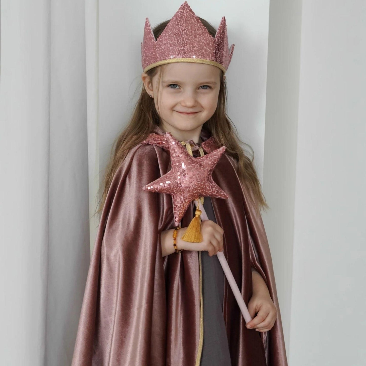 Child in a fairy costume with a pink sequin crown, cape, and wand against a white curtain.